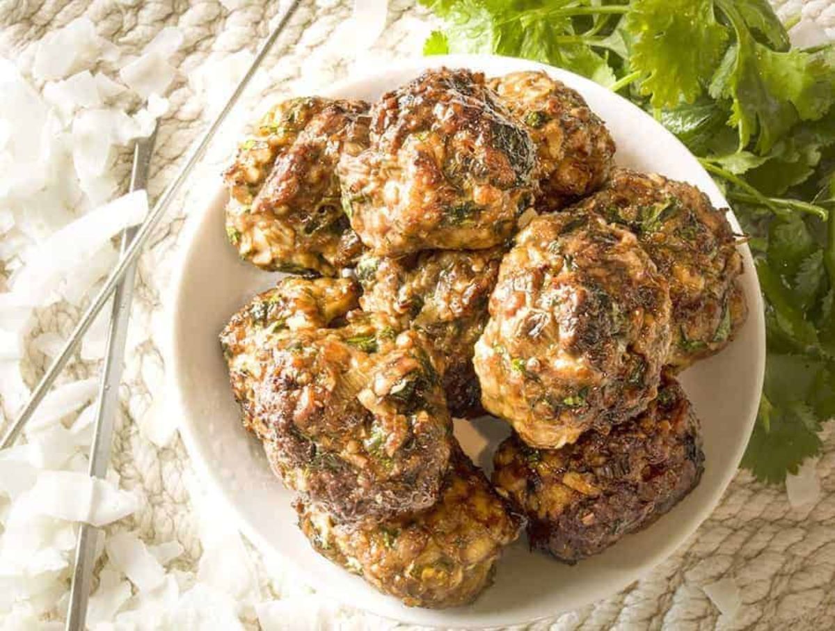 On a cream mat is a white bowl filled with meatballs. TO the right is a bunch of cilantro and to the left is some cutlery