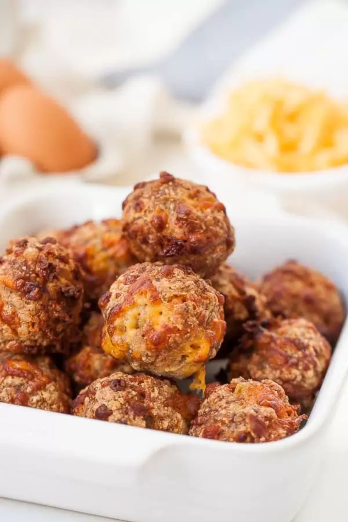 A rectangular oval baking dish is filled with baked sausage balls. In the bacground are blurred pots of toppings and some eggs