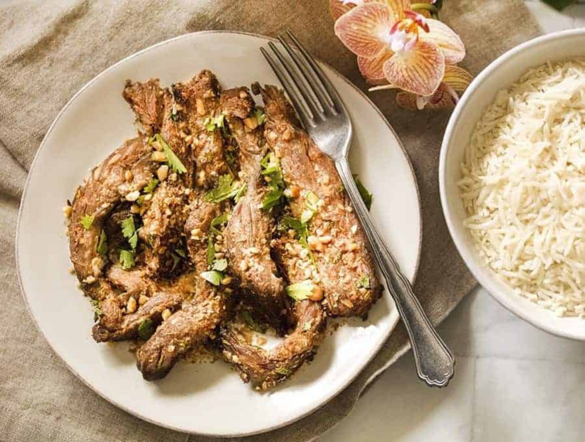 On a cream mat is a white plate with beef satay on it. A silver fork is to the right, with a bowl of rice and a flower