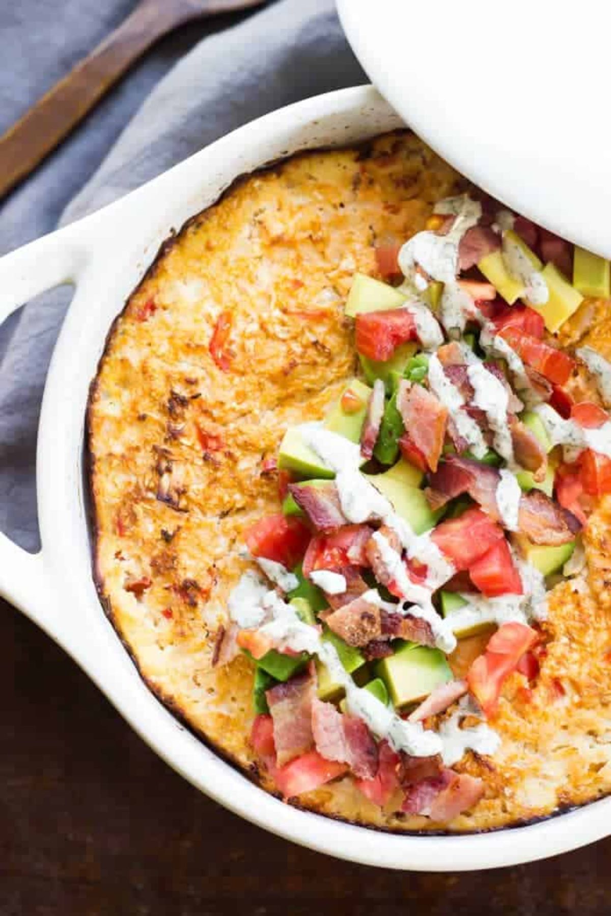 on a pale blue cloth is a white casserole dish, with the lid partially covering it. Inside is a cheesey casserole topped with chopped bacon, avocado and tomato