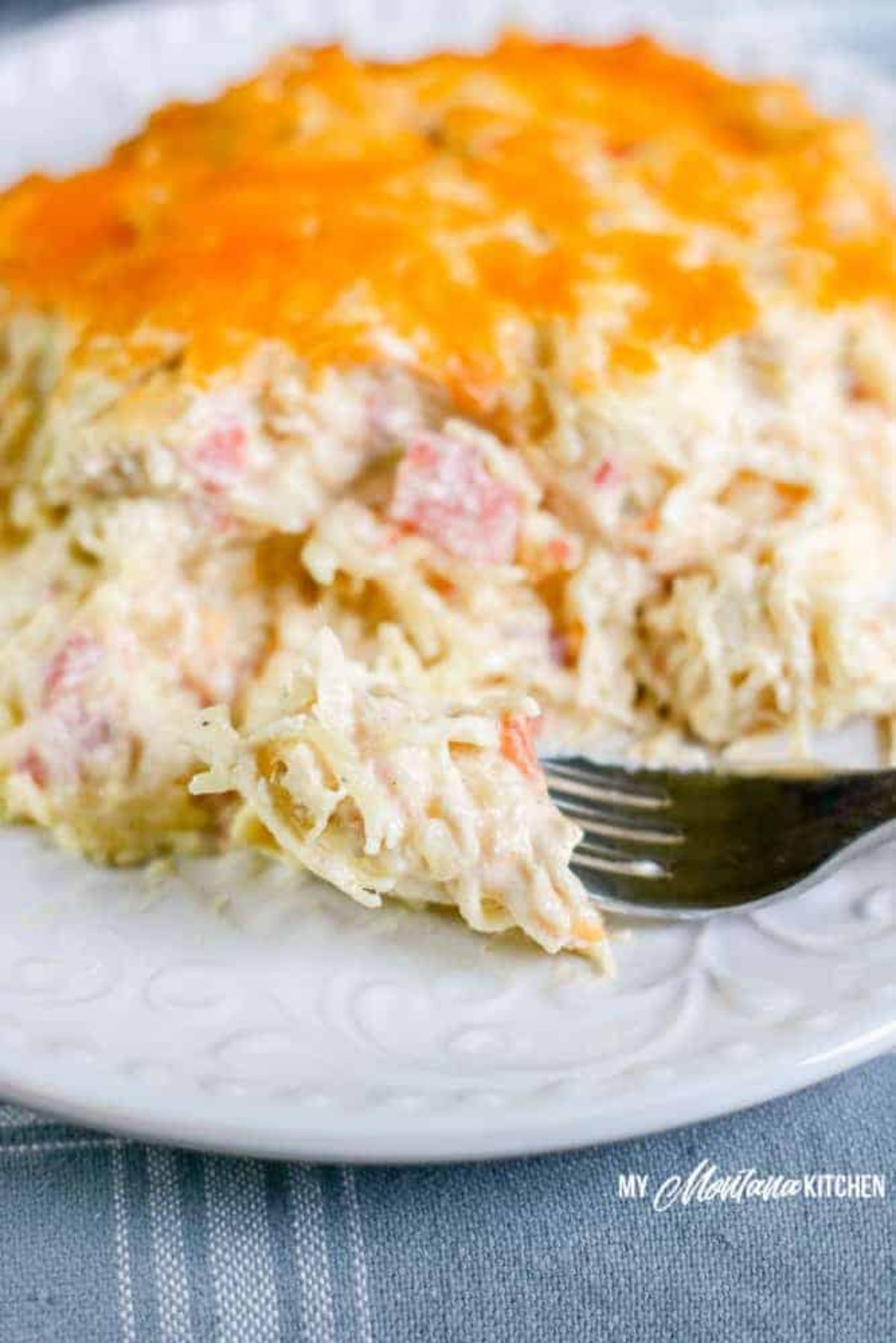 a partially blurred white plate with a cheesy casserole on it, and a metal fork sticking into the portion