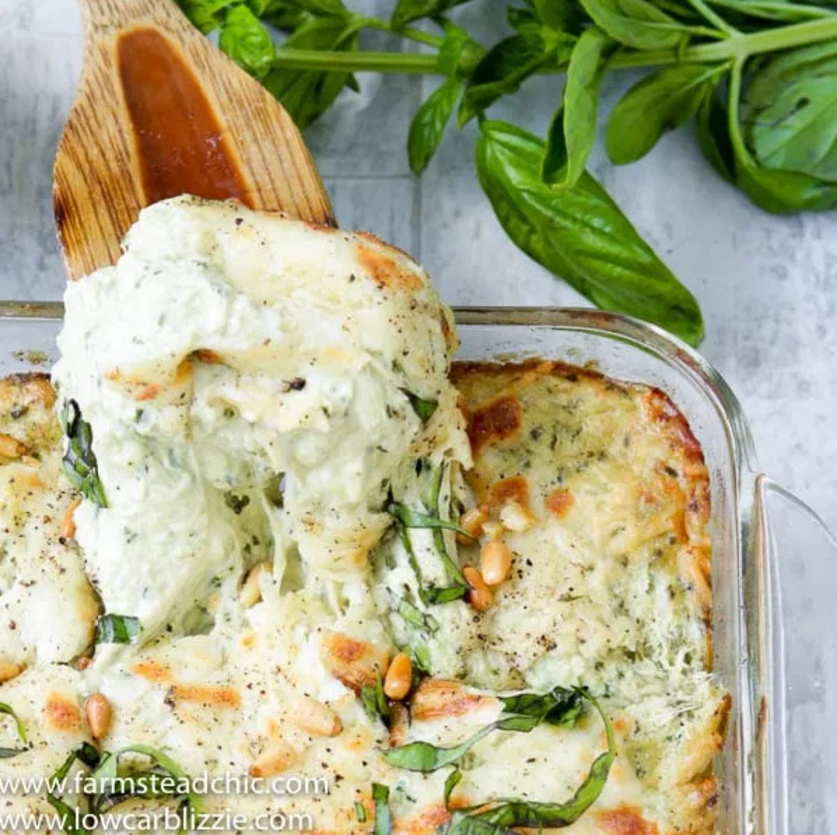 on a white tiled surface, a sprig of basil is at the top of the photo. A wooden spoon sticks into a glass casserol dish full of cheesy casserole with pine nut and basil garnish
