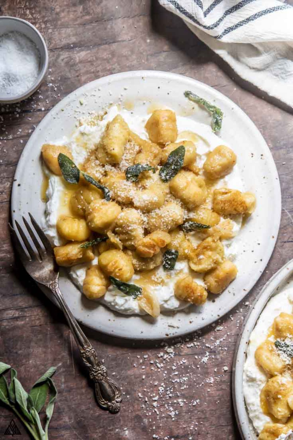 On a dark wooden table is a white plate. On the plate is gnocchi in a creamy sauce with basil leaves. A metal fork is by the side of the plate and a towel sits above it