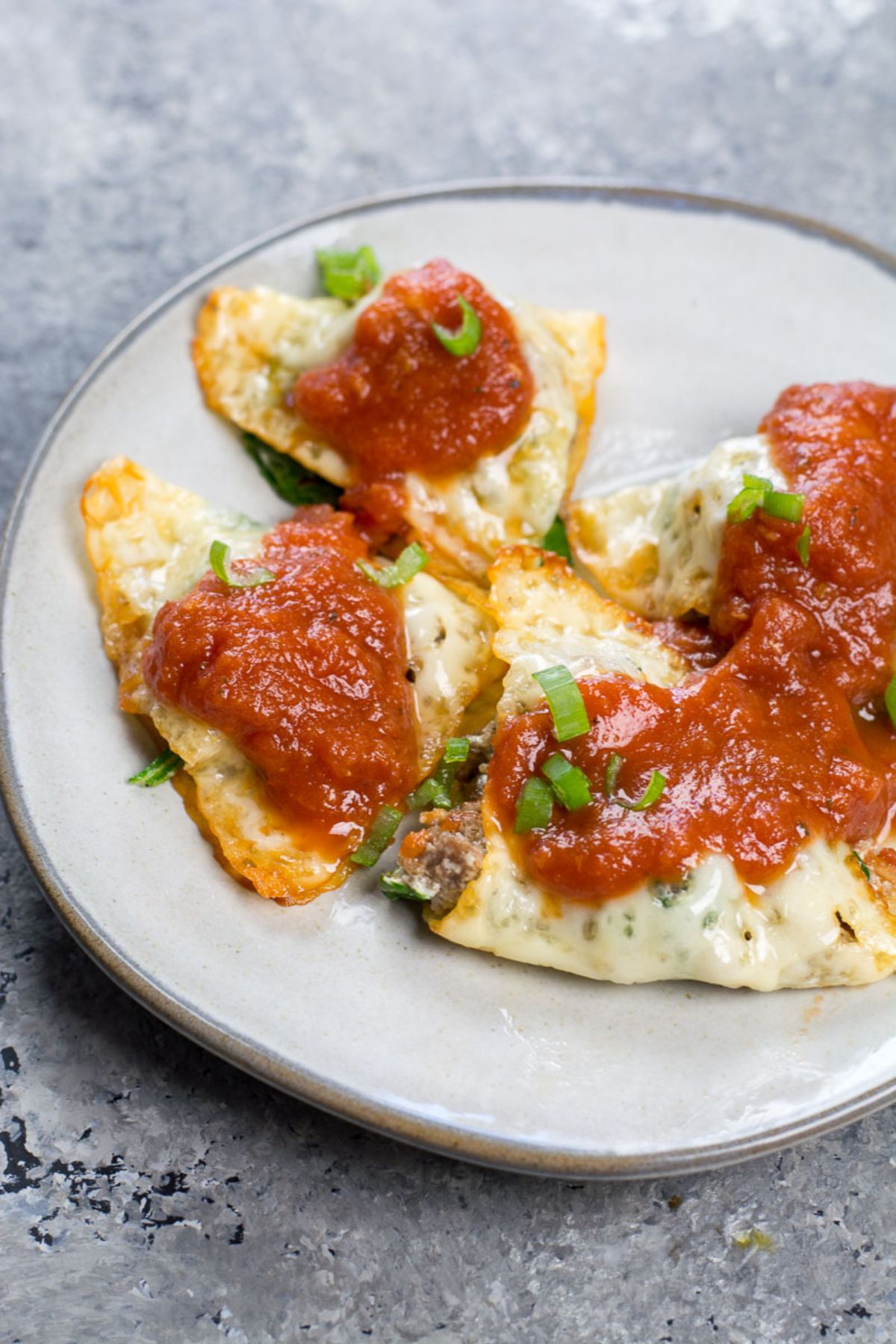 on a gray surface is a small white plate with ravioli. On top is drizzled tomato sauce and chopped chives