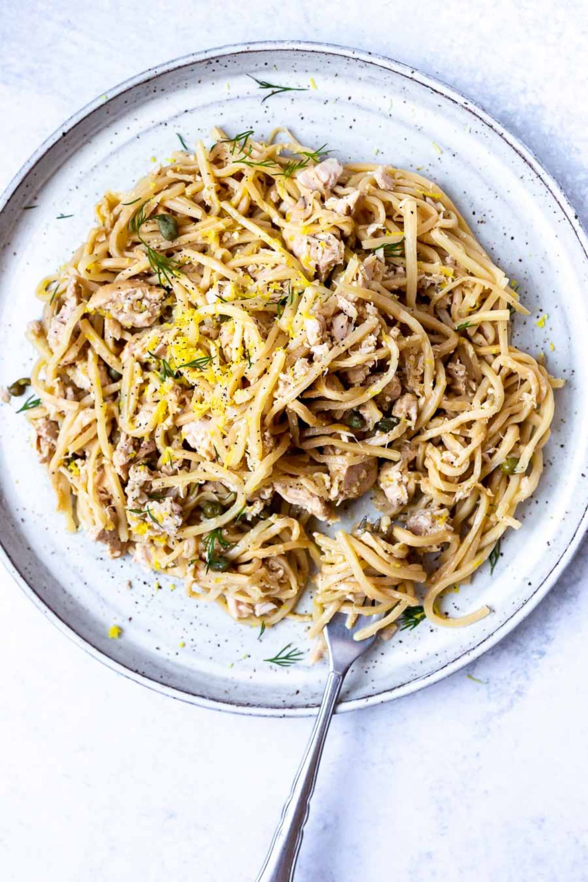 On a white surface is a speckled pottery plate. On the plate is a pile of spaghetti sprinkled with dill, lemon zest and cheese. A metal fork is seen at the bottom