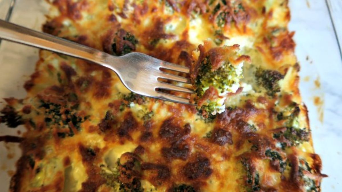 A glass dish holds a cheesy broccoli casserole with darkened top. A fork lifts some of the casserole out of the dish