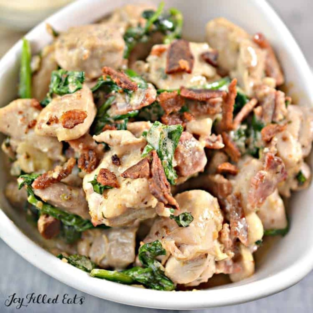 The text reads "Joy Filled Eats". In a white bowl is a pile of chicken in creamy sauce with cripsy bacon bits and green leaves wilted through