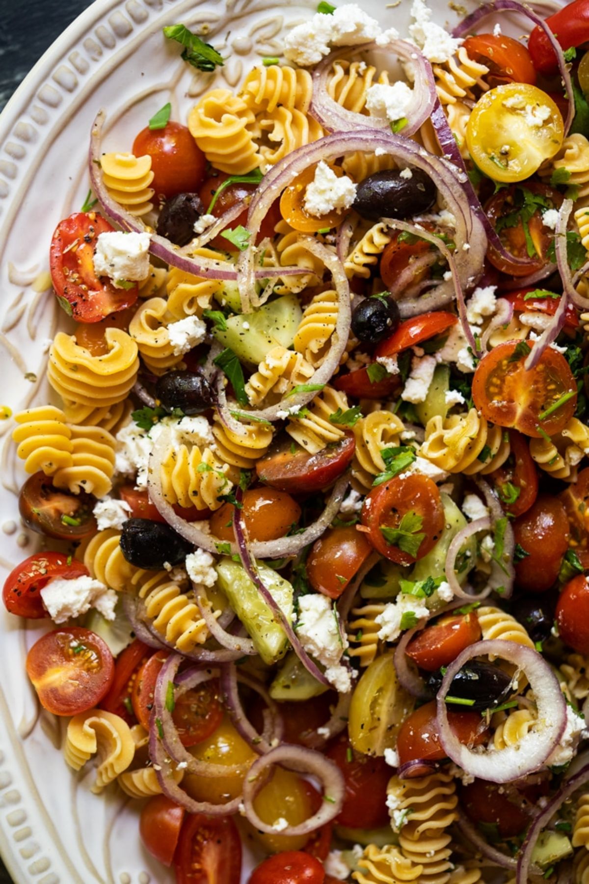 A partial shot of a white plate of spiralled pasta shapes mixed through with cherry tomatoes, red onion rings, cucumber and crumbled feta cheese