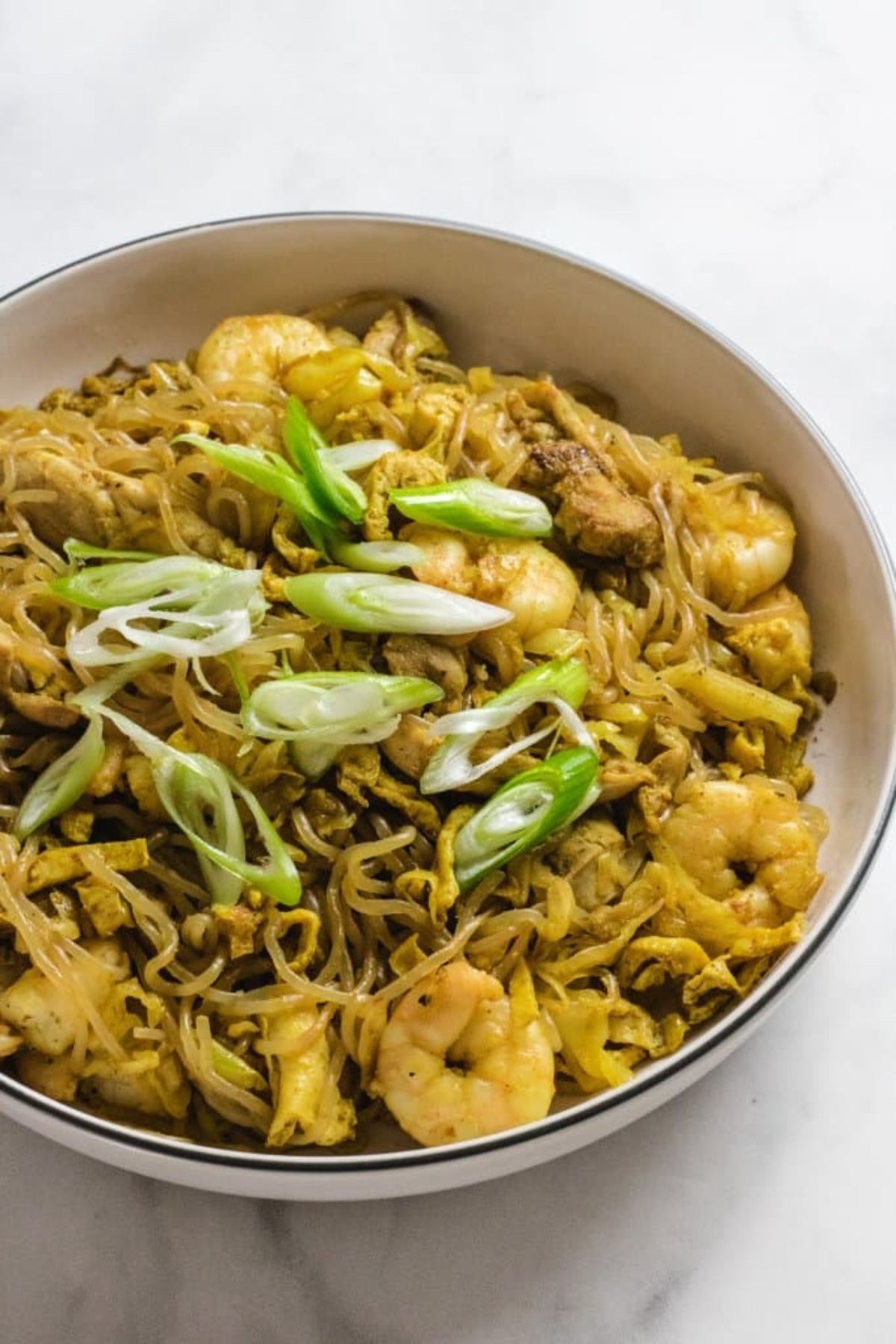 on a white surface is a white bowl with a blue rim. Inside is a pile of spaghetti with small peeled prawns, chopped spring onions and a yellowish sauce