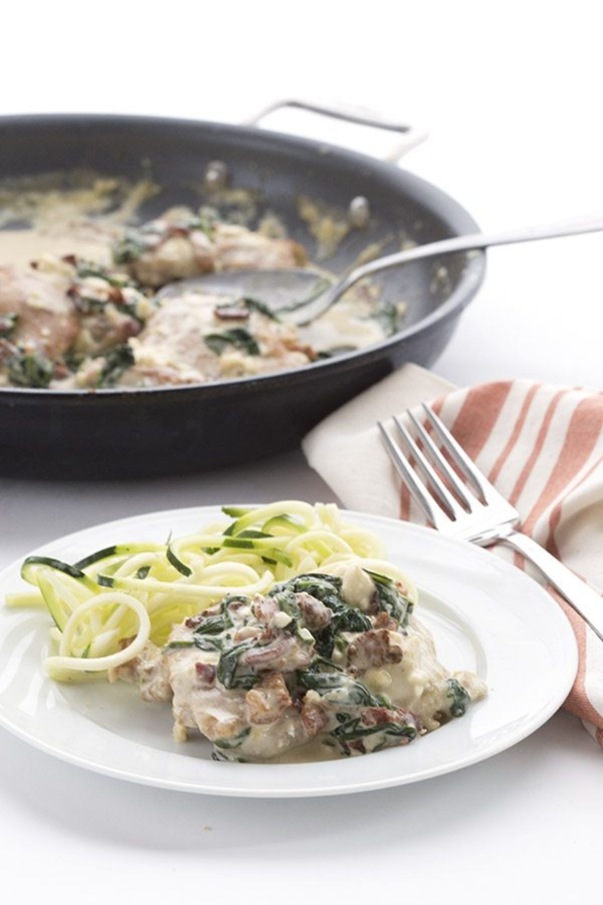 a white ocuntertop on which sits a dark gray skillet containing meat in a creamy sauce. In teh foregraound is a white plate with some of that meat and sauce, green wilted leaves and some spiralized courgette