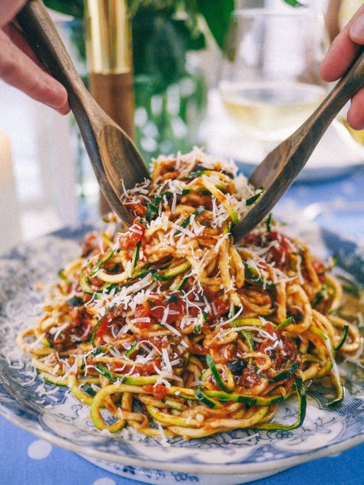 on a light blue tablecloth with white spots is a delft style dish. On top of it is some spiralized courgette covered in tomato sauce and parmesan cheese. Two wooden serving spoons are being hed by two hands digging in to the pasta