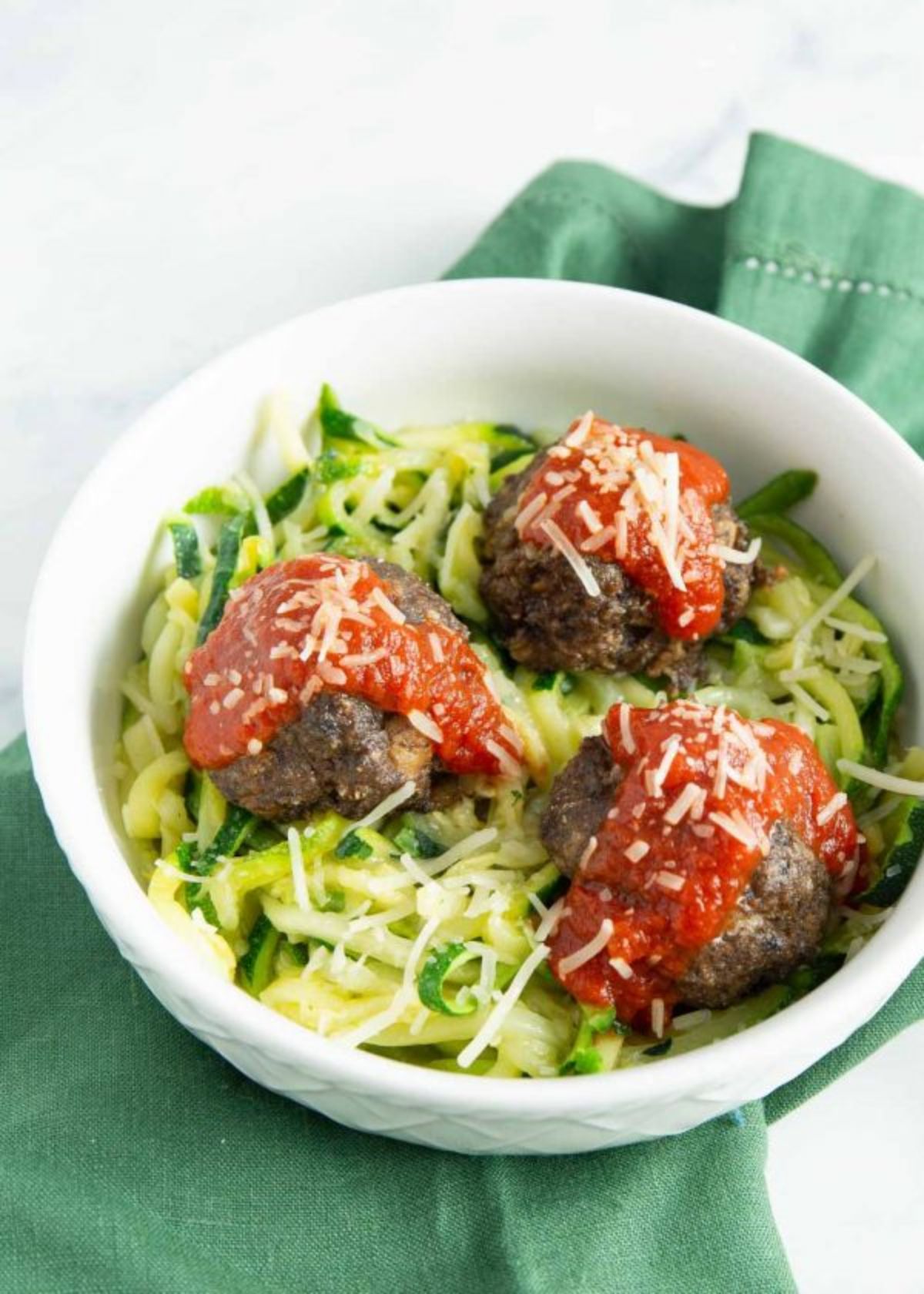 a white surface with a green cloth on top. On top of this is a white bowl filled with courgetti, 3 large meatbalss and 3 blobs of tomato sauce