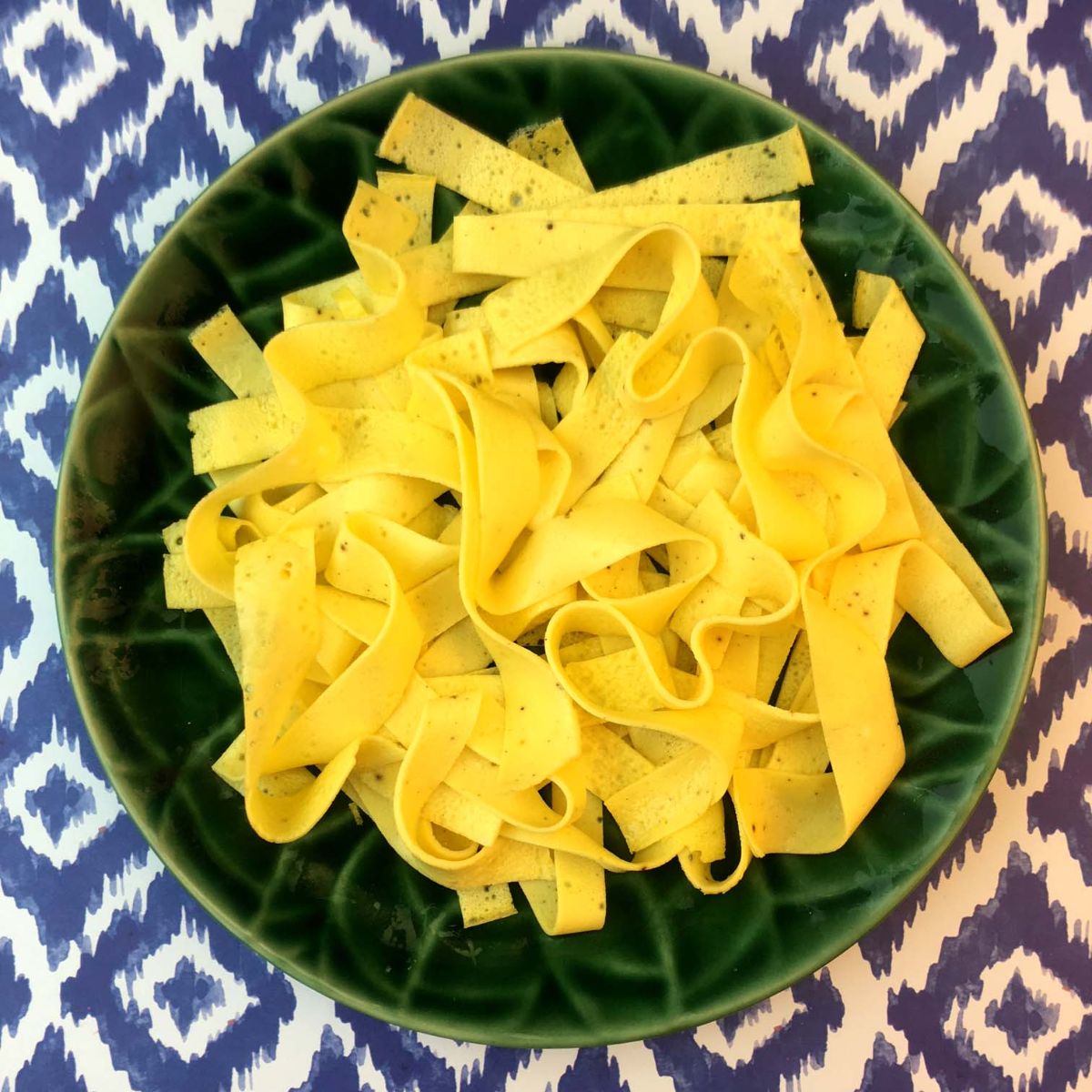 a blue patterened surface iwth a green bowl on top. Inside is a pile of egg noodles