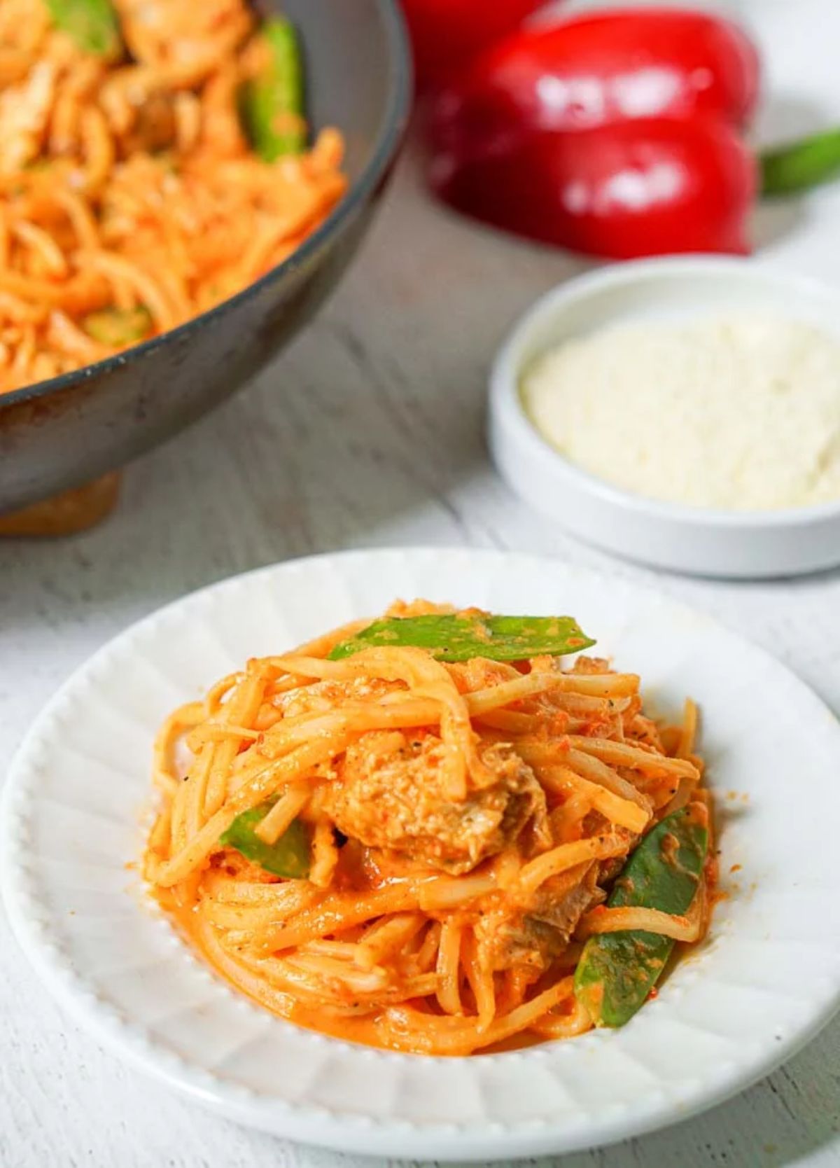 a partial shot of a dark skillet with pasta covered in tomato sauce in it. To the right of the photo is half a red pepper and a dish of grated parmesan cheese. In the foreground is a white plate filled with the pasta dish