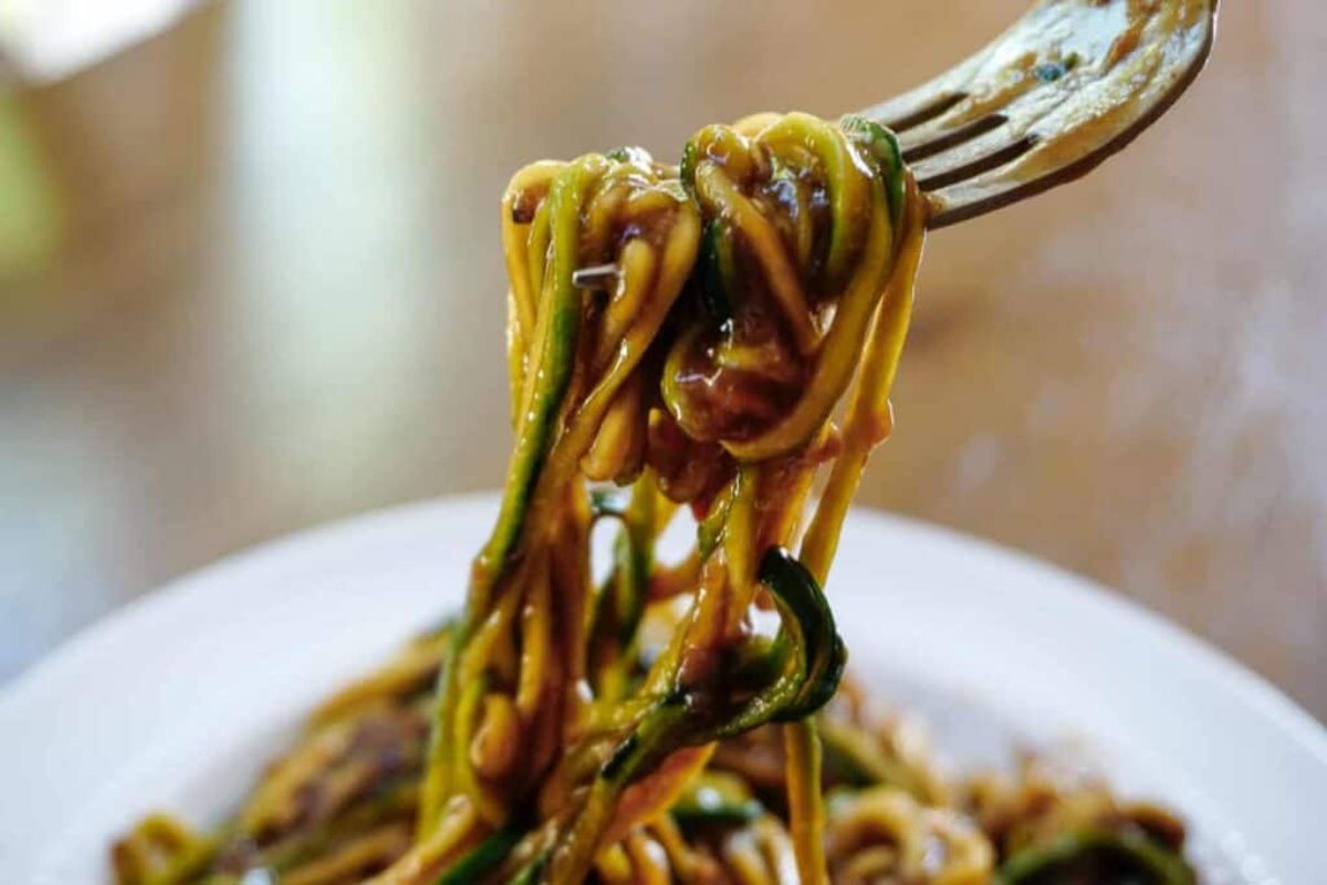 A partial shot of a white bowl of courgetti covered in a gravy-like sauce. A metal fork is pulling some of the noodles upwards