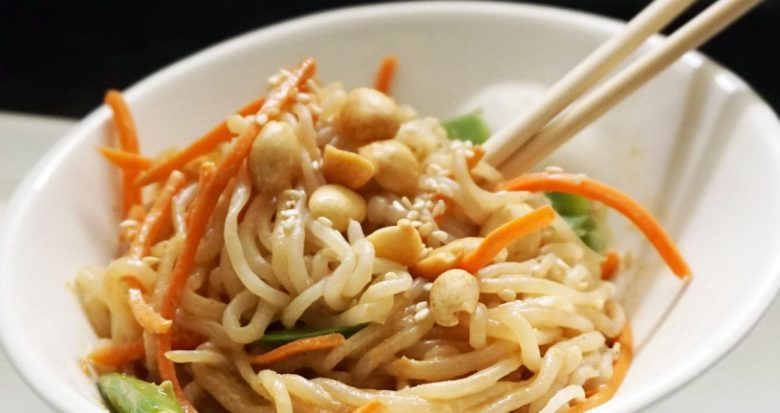 a white bowl with pale noodles mixed with matchstick carrots and leaves. Cashews are scattered on top and two chopsticks are sticking out of the dish