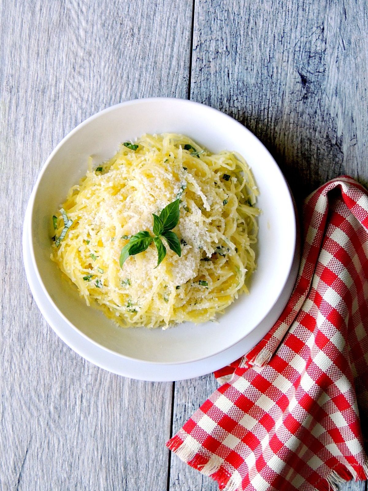 a whitewashed table with a red and white checked cloth in the front right of the frame. In the ceter is a white bowl with spaghetti sprinkled with parmesan cheese and basil leaves