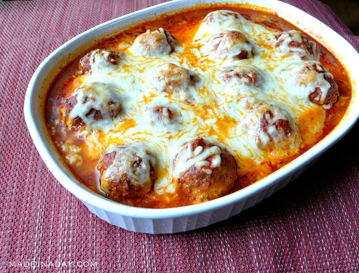 on a red tablecloth is a rectangular casserole dish filled with baked meatballs in a tomato sauce and topped with cheese