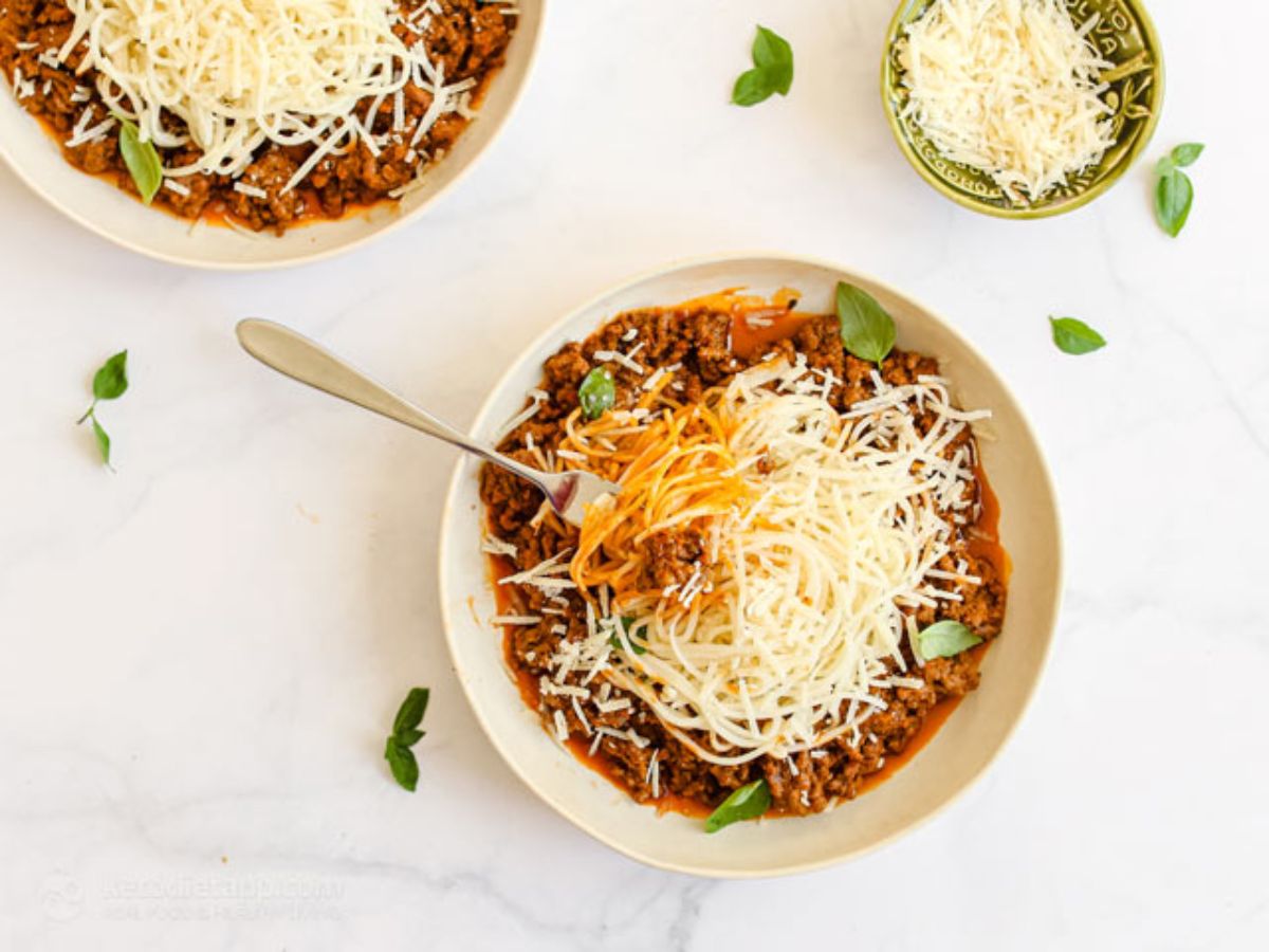 two large bowls on a white surface filled with a meat and tomato sauce and topped with cheese. On esmall bowl sits to the right filled with grated cheese. Basil leaves are scattered over the frame