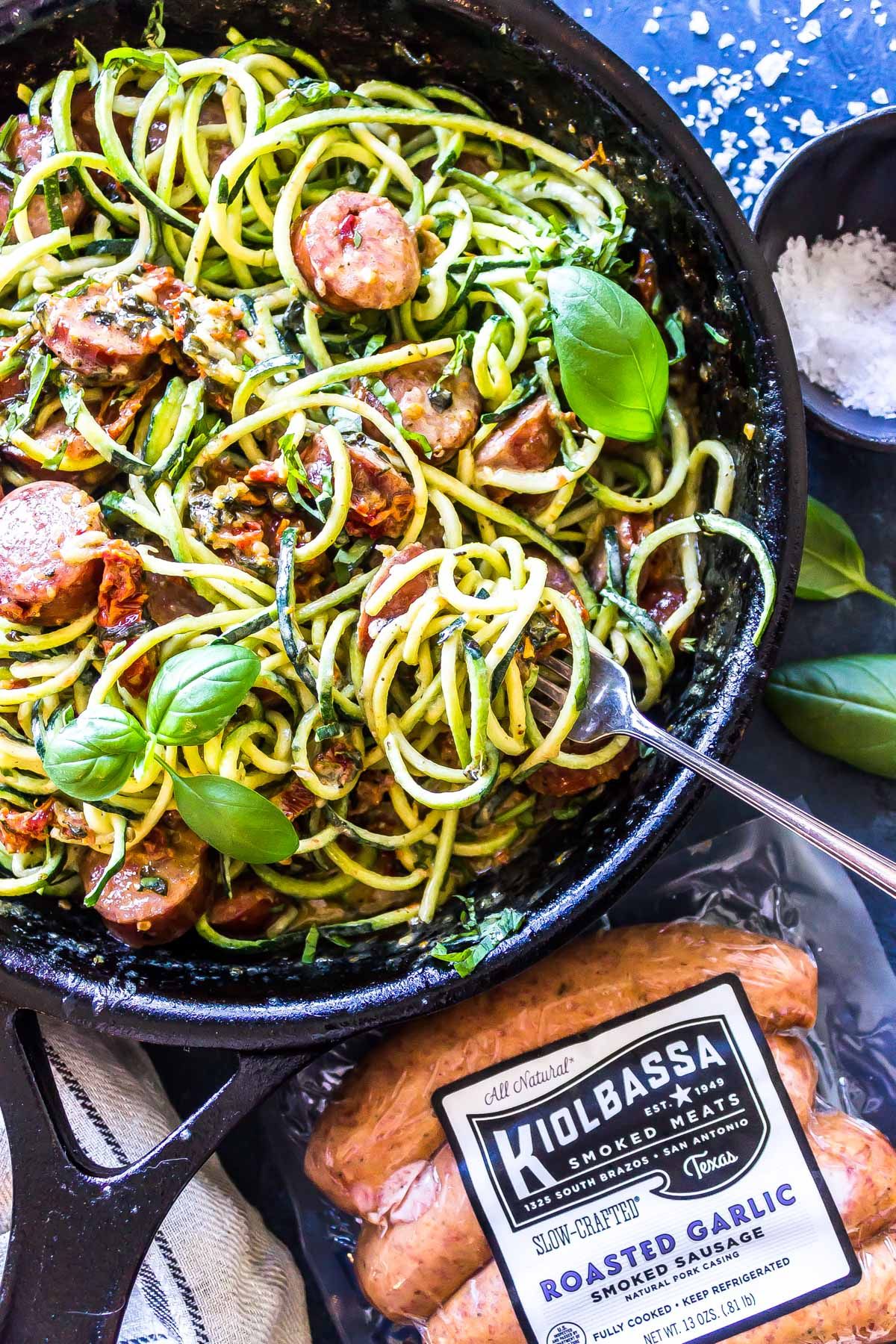 a black skillet contains courgetti mixed with sliced sausage. In the foreground is a packet of the Kiolbassa Roasted Garlic smoked sausage