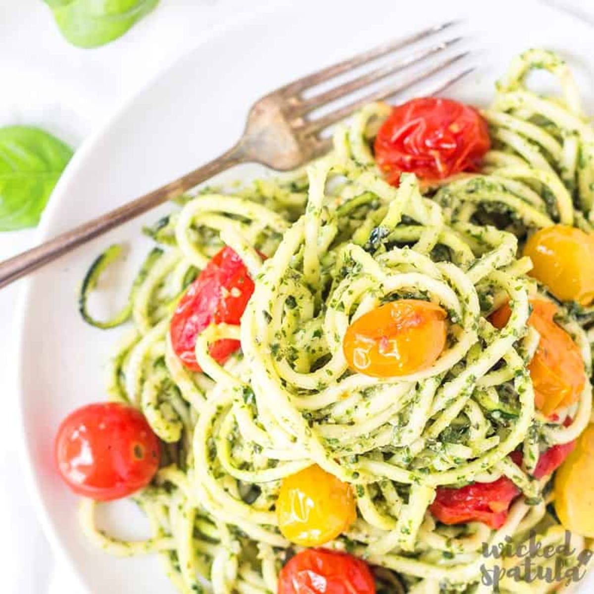 A partial shot of a white bowl filled with spaghetti mixed with pesto sauce and cherry tomatoes. A metal fork is at the top of the bowl