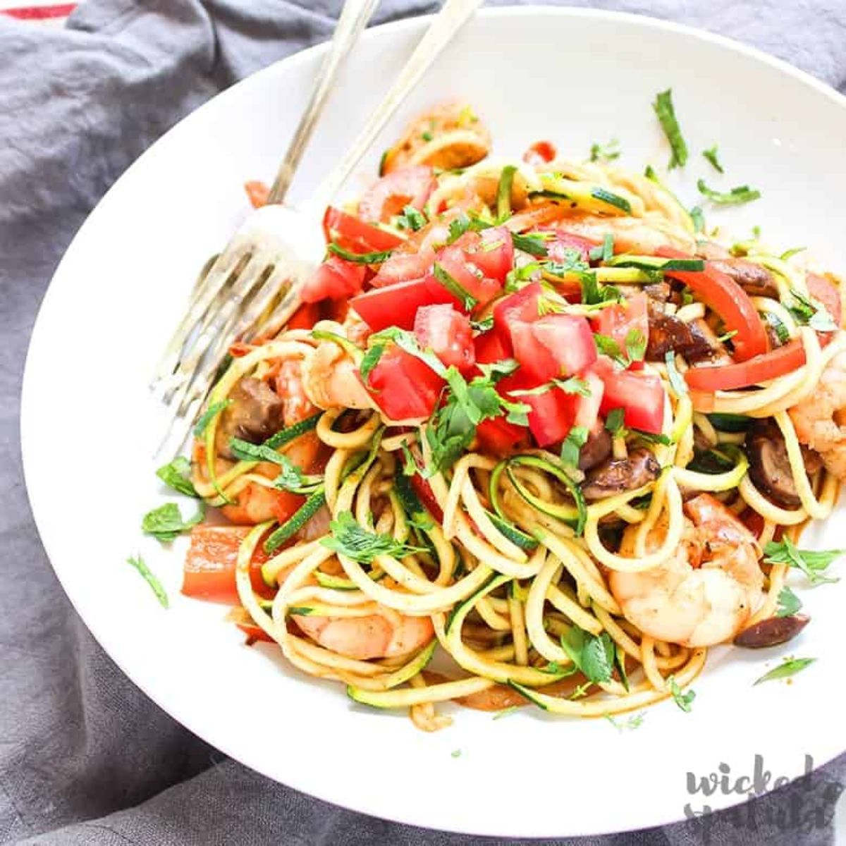 on a rumpled gray cloth is a white bowl filled with courgetti, mixed with proawns, chopped tomatoes and basil