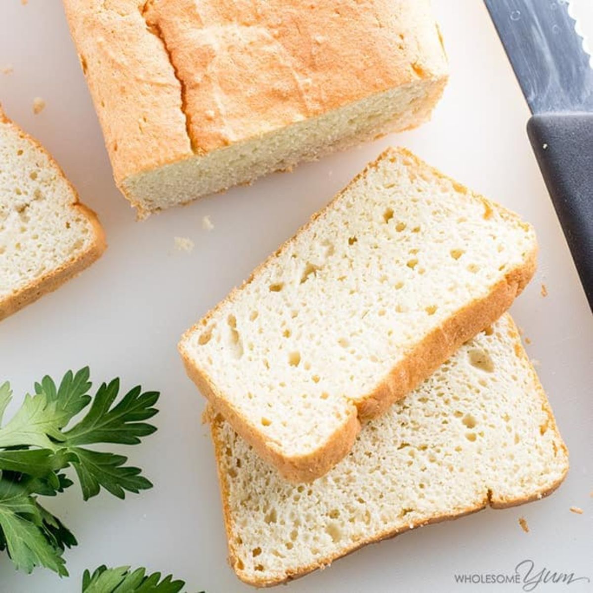 On a white surface is a partial shot of a rectangular loaf of white bread, two cut slices are in the front of the shot on top of each other. Parsely is on the left of the shot