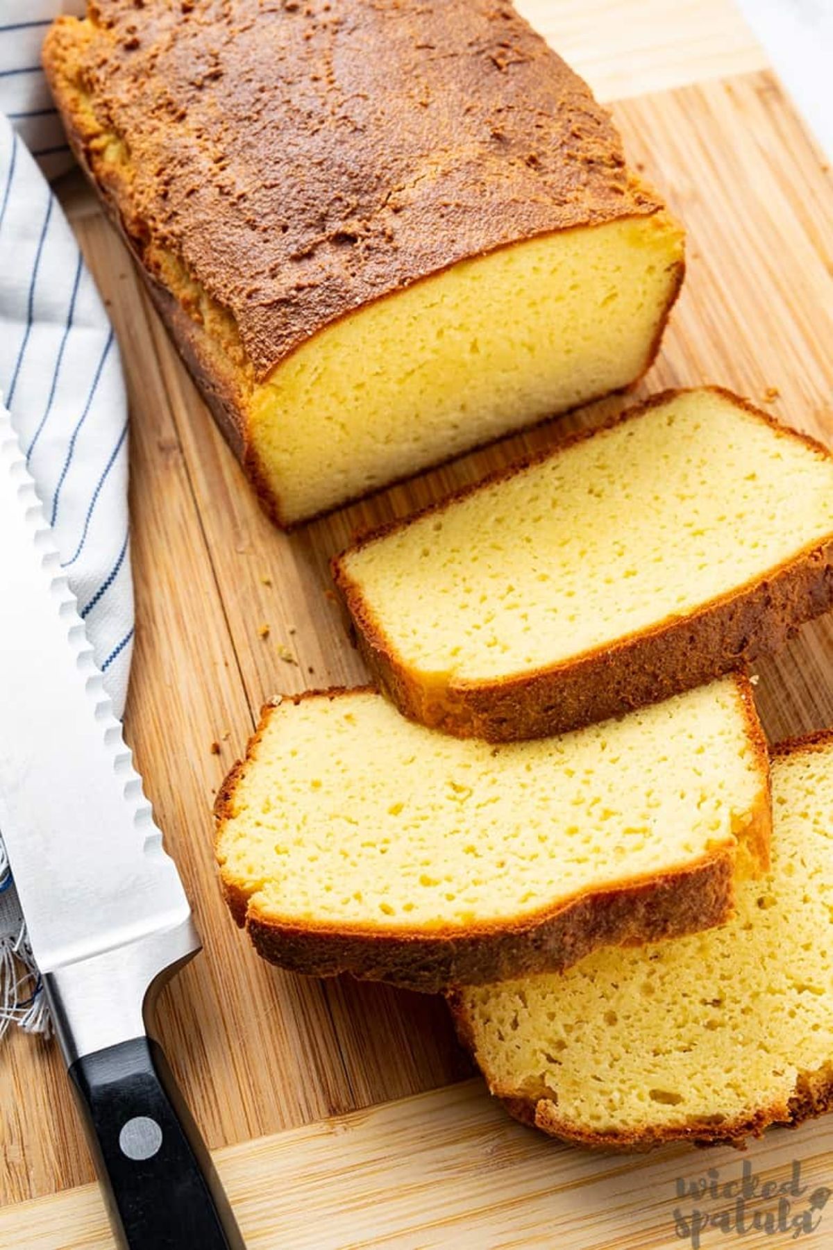 On a woodean counter is a yellow loaf of rectangular bread, with 3 slices cut out of it stacked on each other. On the left is a striped cloth with a bread knife on top of it