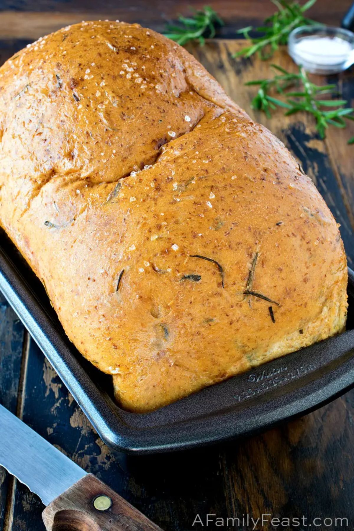 On a wooden kitchen table is a loaf tin with a risen loaf od bread in it. Bits of rosemary can be seen within the loaf. Rosemary sprigs are scattered on the top right and a knife can be seen on the bottom left