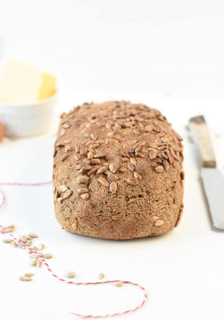 a white background is a seeded square loaf of bread. Seeds are scattered to the left, and a bread knife is on the right. In the background can be seen a blurred pot of butter