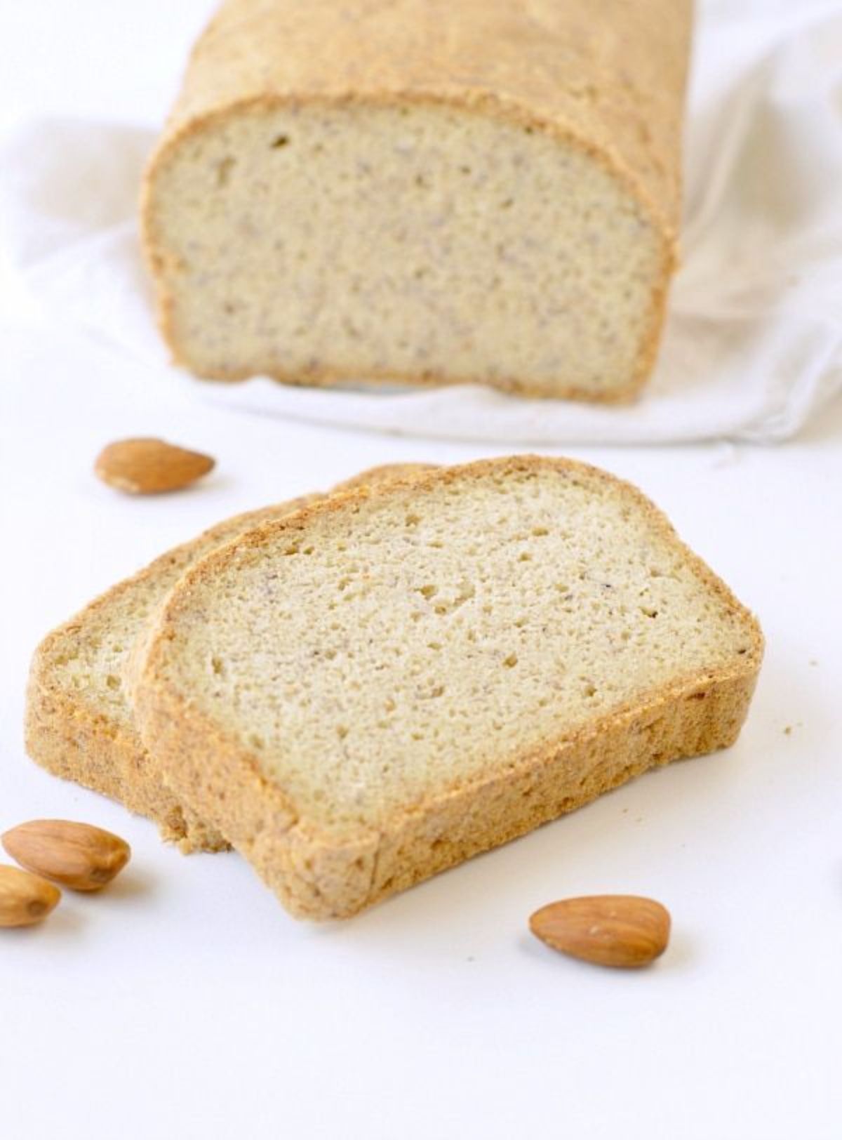 ON a white cloth is a cut loaf of bread. In front is two slices ofe bread stacked on each other and almosnds are scattered around