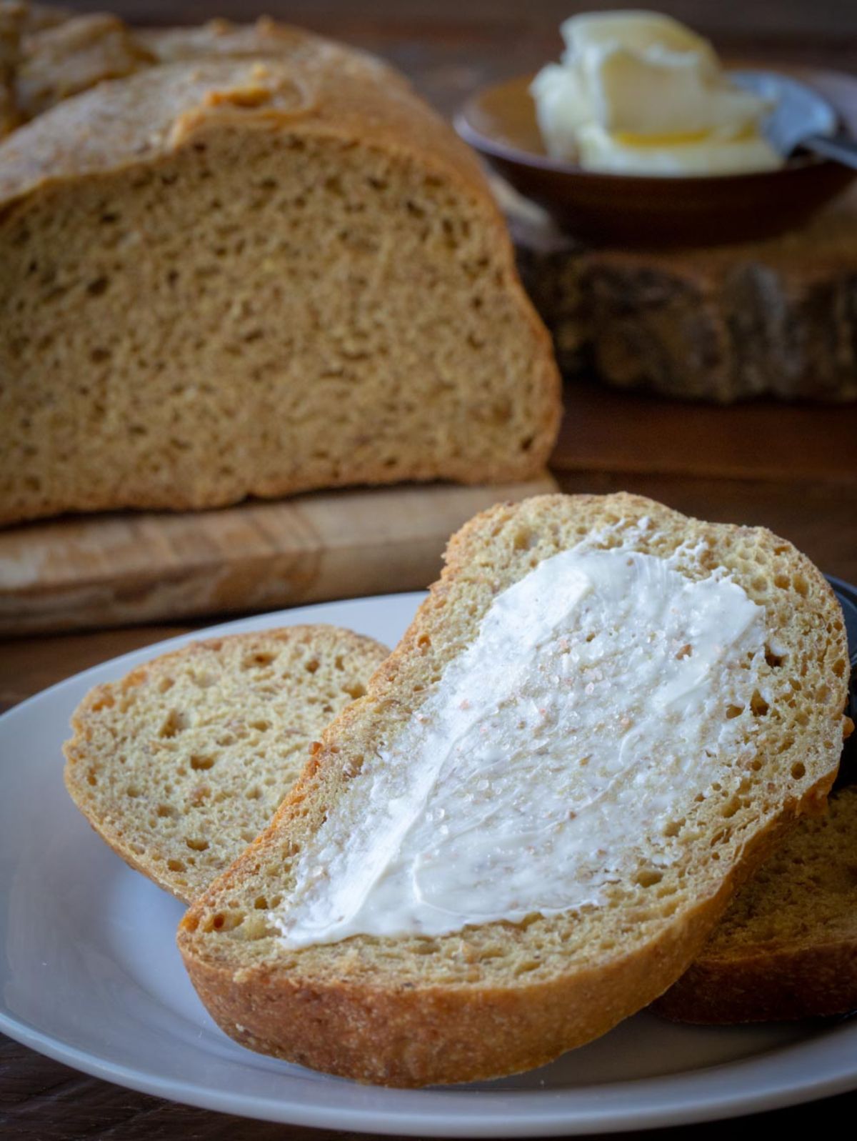 On a white are two slices of bread. THe top one has butter spread on it. In the background is the rest of the loaf on a wooden board.