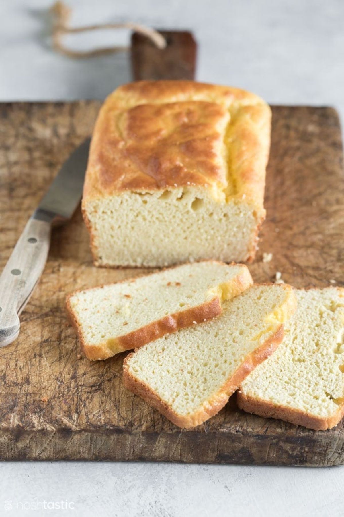 On a wooden board is a pale rectabgular loaf of bread with 3 slices cut and laid in front of it. On the left is a knife.