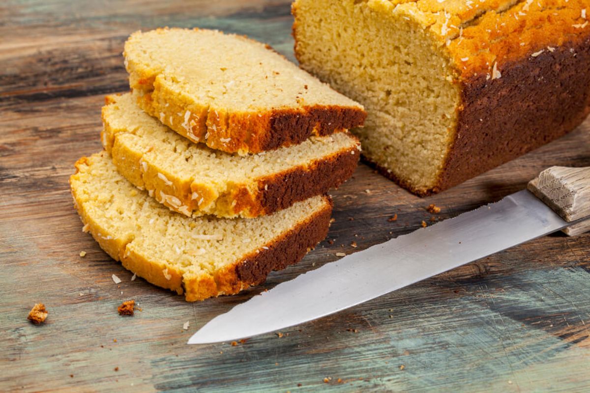 On a wooden table is a rectangular loaf of bread with a yellow tinge. 3 slices are laid on top of each other and a bread knife is in the front of the shot