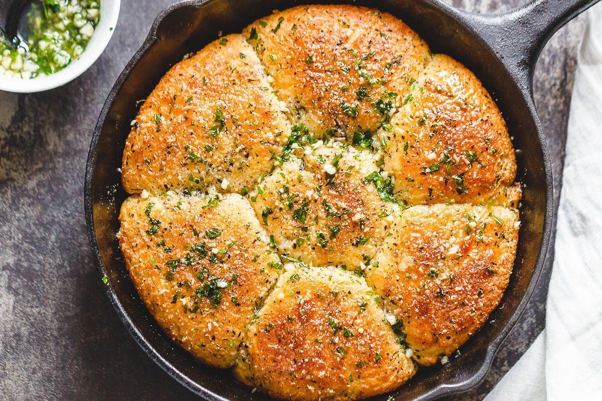 On a gray stone surface is a cast iron skillet with a round loaf in it, sprinkled with herbs. The loaf is portioned into 6 to pull apart