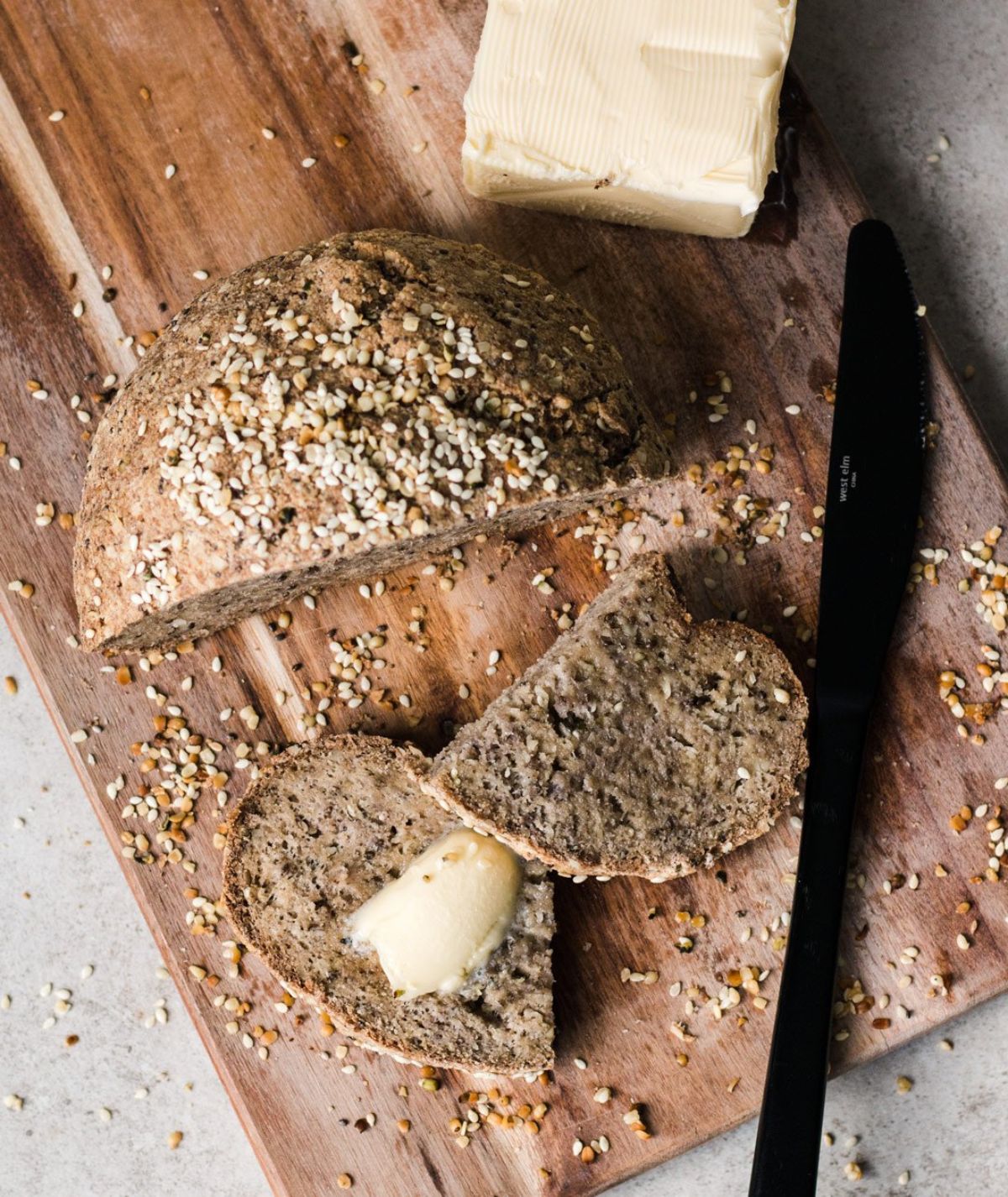 A wooden board is on a light gray counter. On the board is a block of butter, half a round loaf of bread, and a black knife. One slice of the bread is cut in half and butter has been spread on it