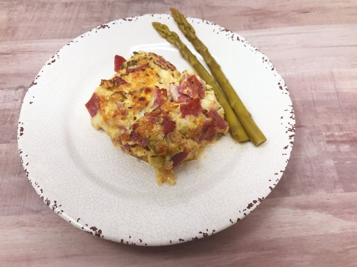 On a white, speckled plate is a square portion of a ham and cheese sauce casserole. Next to the portion are two pieces of asparagus