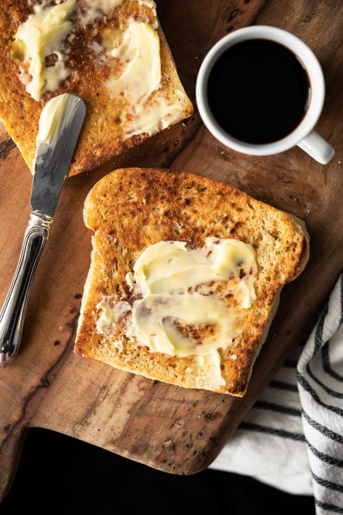 On a wooden board are 2 slices of bread with butter spread on them. A butter knife rests on one of the slices and a white cup of black coffee is at the top