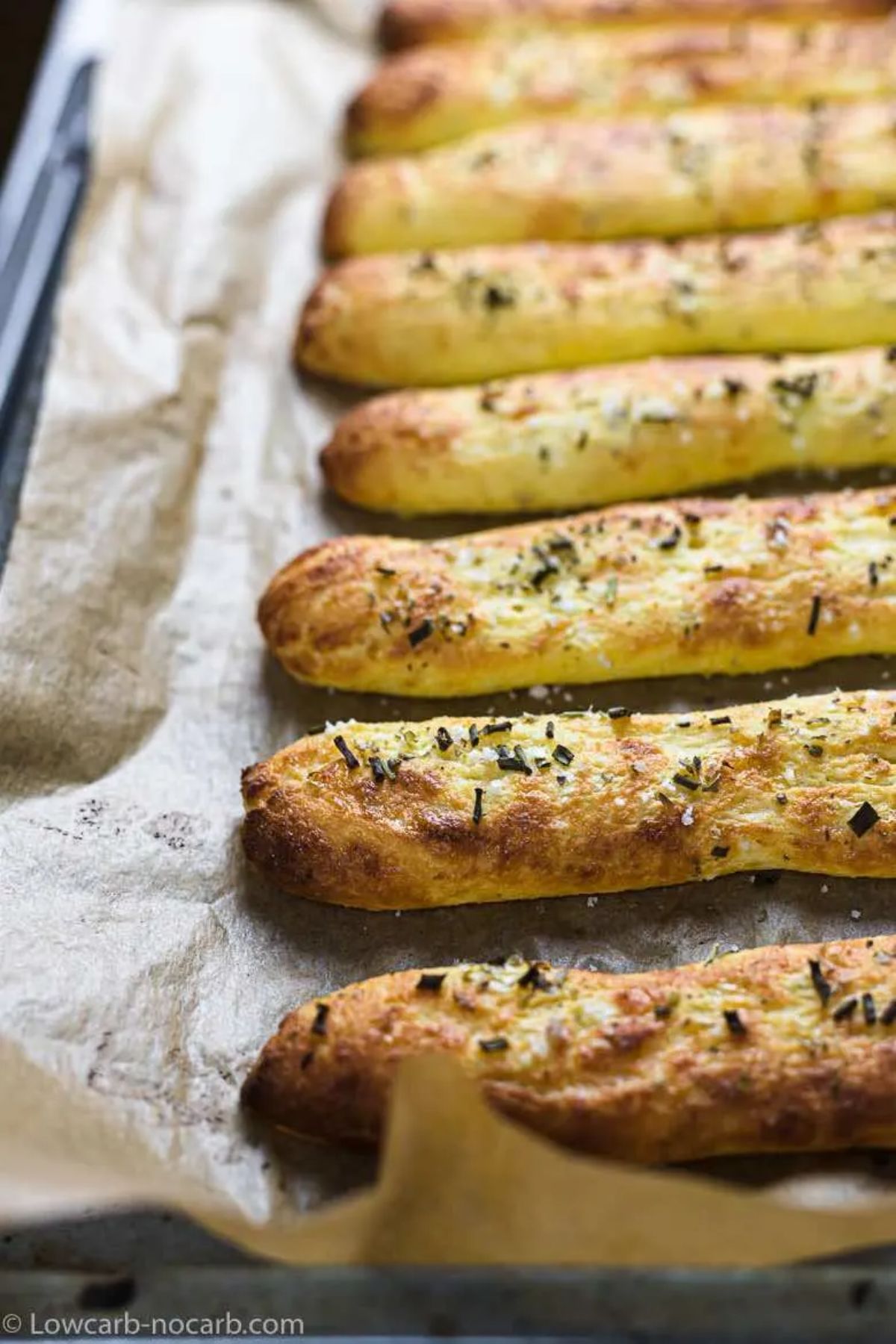 On a baking tray and sheet of baking paper are 8 long sticks of bread with herbs sprinkled on top