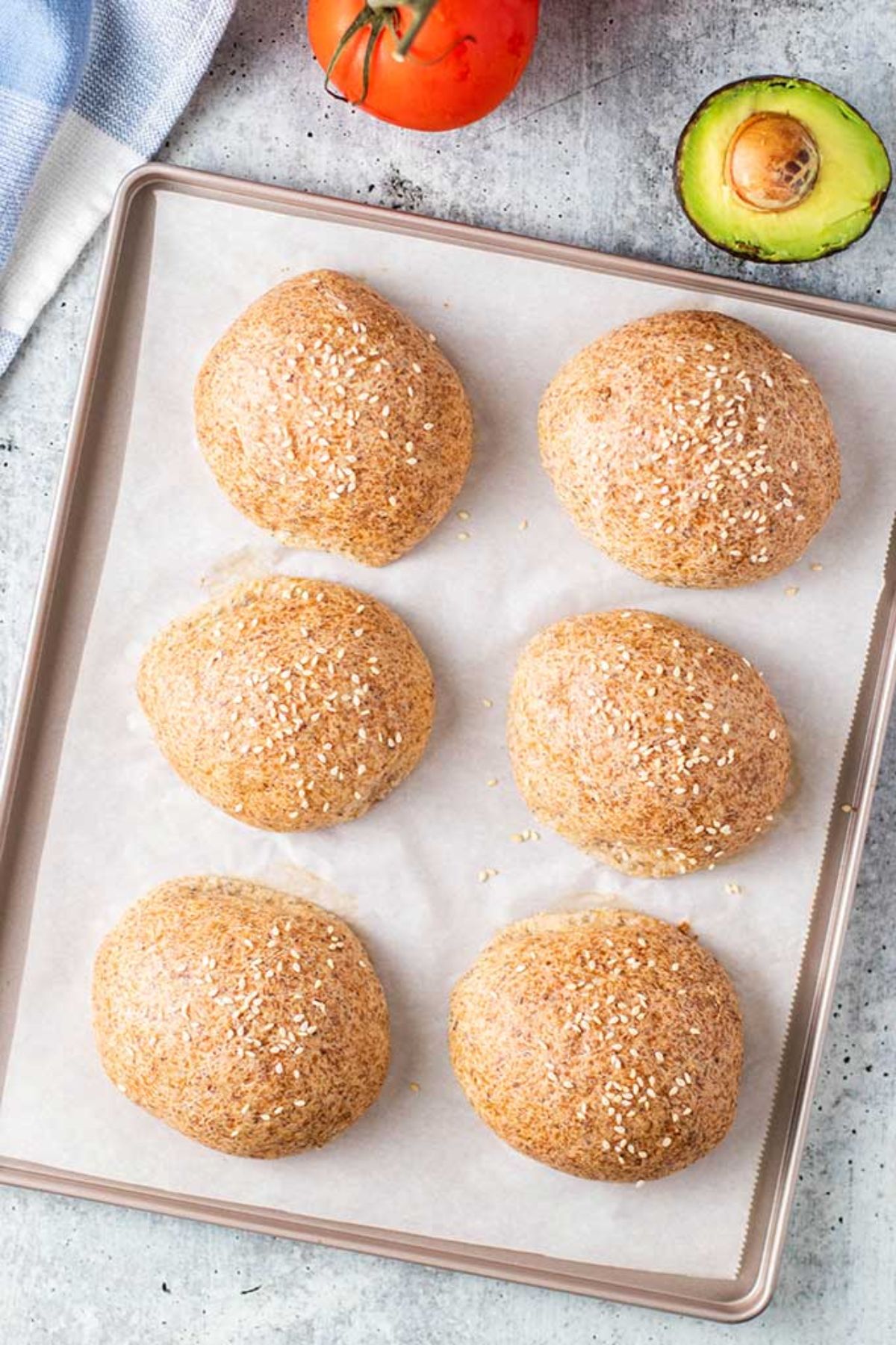 A birds-eye shot of a baking tray lined with paper and 6 brown round bread rols on it. On the top of the shot is a tomato and half an avocado