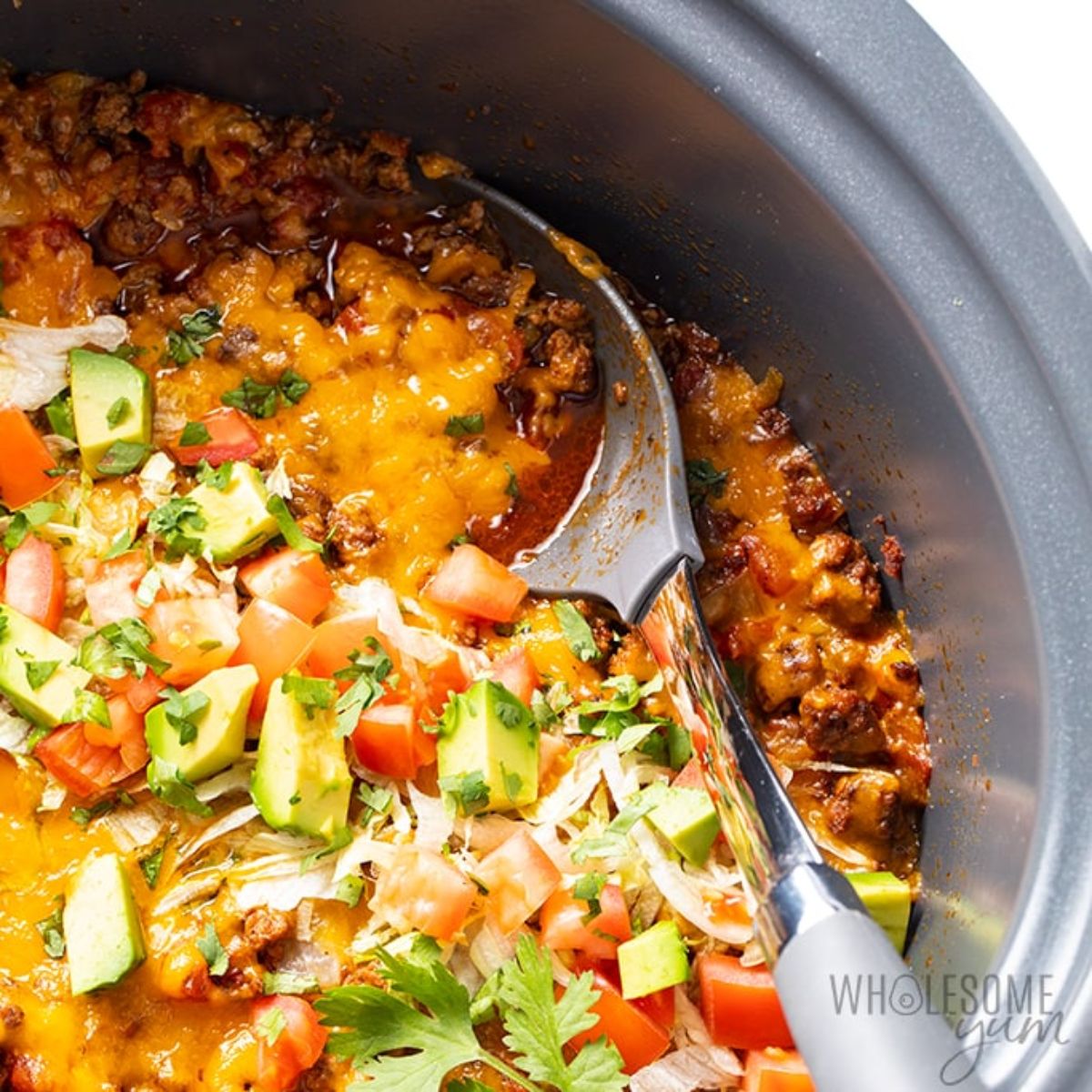 A dark casserole dish contains a casserole with avocado, tomoato and parsley garnish. A silicone spoon sits in the casserole