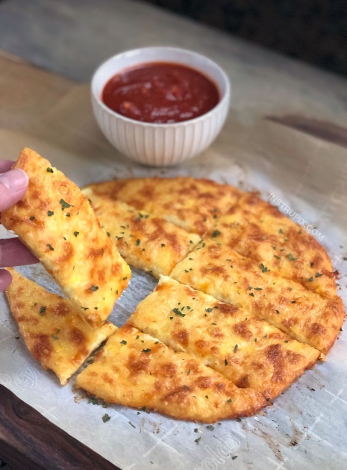 On a sheet of baking paper is a round flatbread cut into strips. An hand is picking up one piece. At the back of the shot is a white bowl if red sauce