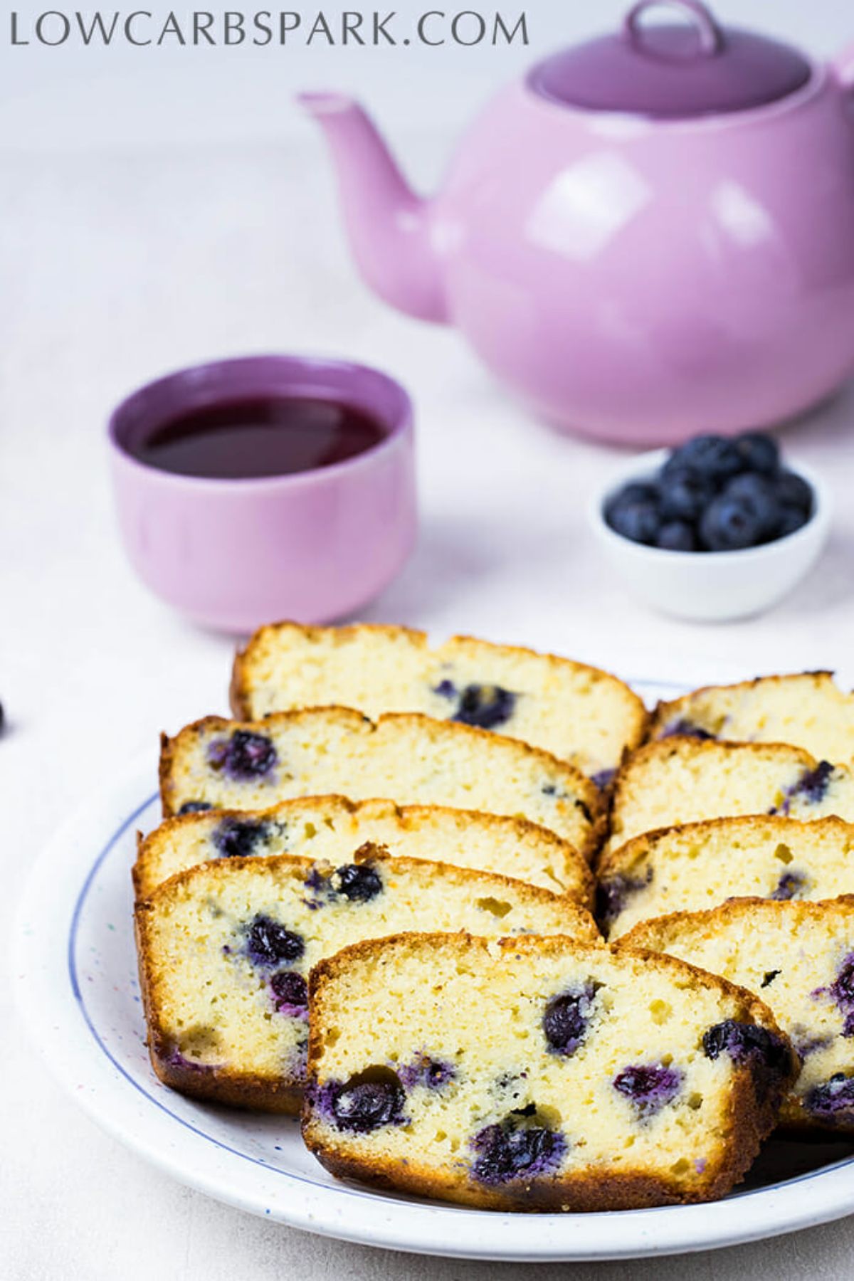 A lilac tea pot and teacup is in the background with a small white bowl of blueberries next to them. In the front is a white plate with a blue rim and stacked slices of blueberry loaf on it