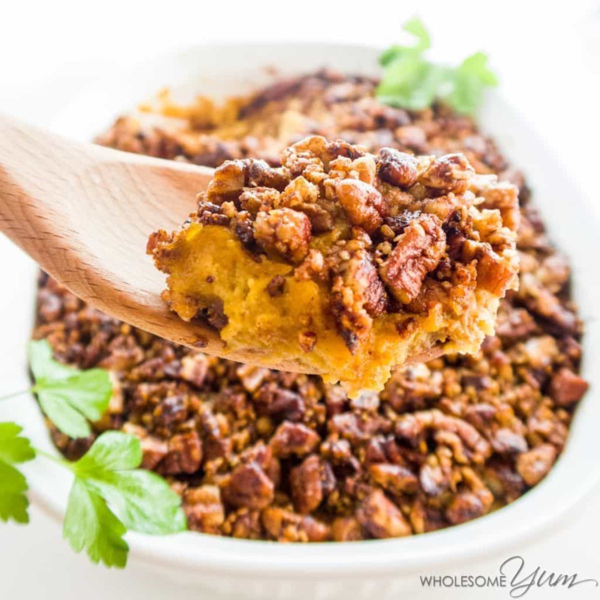 A white bowl of sweet potato casserole with parsely garnish is in the background. In the foreground is a wooden spoon with a spoonfull of the casserole on it